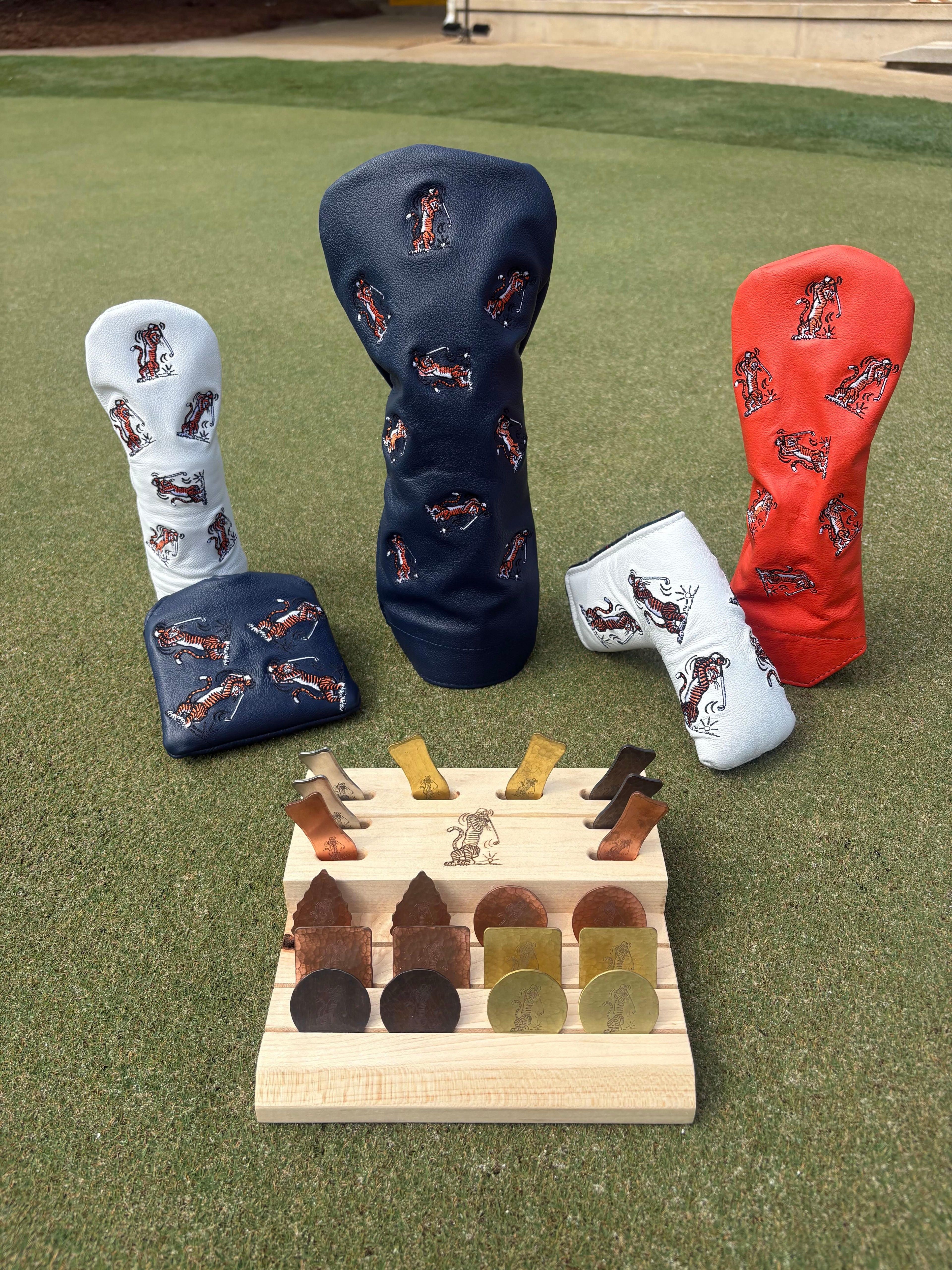 Four golf club headcovers in navy, white, and red with a horse logo, displayed on a green grass background alongside a wooden coin and club holder stand.